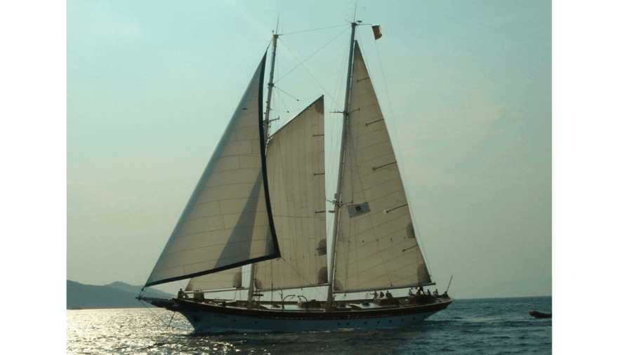 Teambuilding - Groei & Verbinding, Zeilworkshops in Lefkas. Een majestueuze zeilboot met grote witte zeilen glijdt over de oceaan. De vlag wappert boven op de mast tegen een serene, zonovergoten hemel.