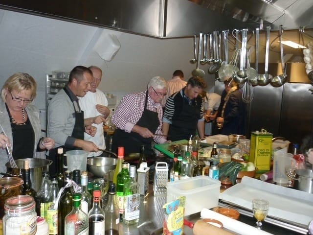 Een drukke keuken met meerdere mensen die samen koken, schorten dragend, omringd door ingrediënten en keukengerei. De sfeer is collaboratief en levendig.