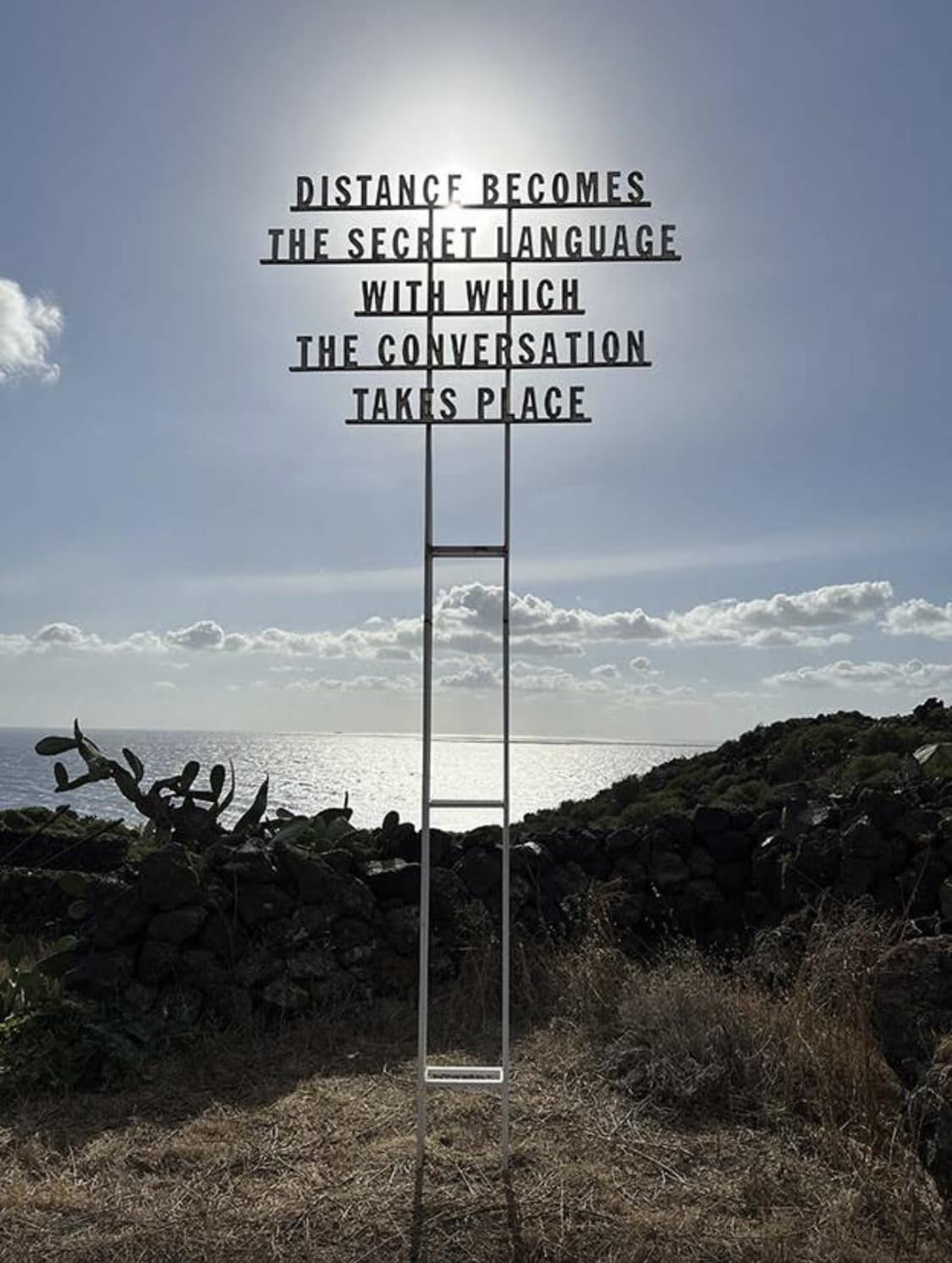 Kunst op Pantelleria. Metalen ladder met woorden tegen een heldere hemel en zee, met de tekst: "Afstand wordt de geheime taal waarin het gesprek plaatsvindt." Rustiek en contemplatief.