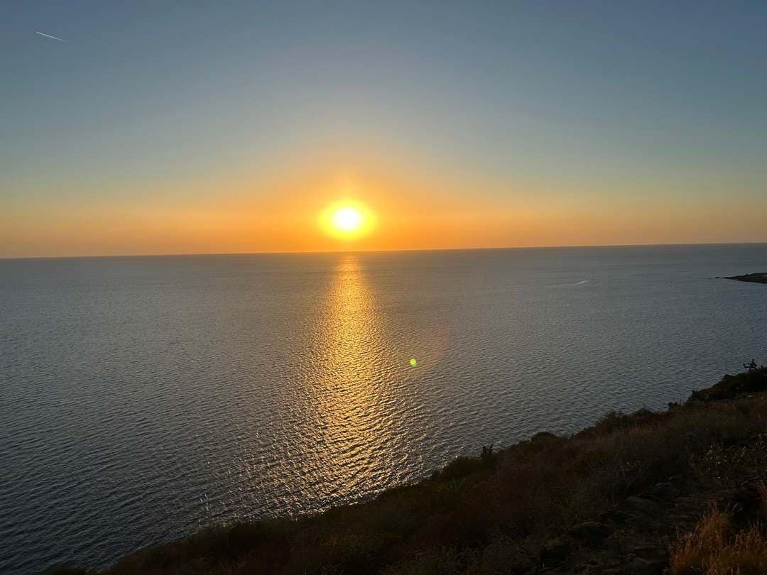 Zonsondergang boven Pantelleria, een uitgestrekte oceaan met een heldere hemel. De zon werpt een gouden weerspiegeling op het water, wat een vredige, serene sfeer creëert. Een rotsachtige kust is op de voorgrond te zien.