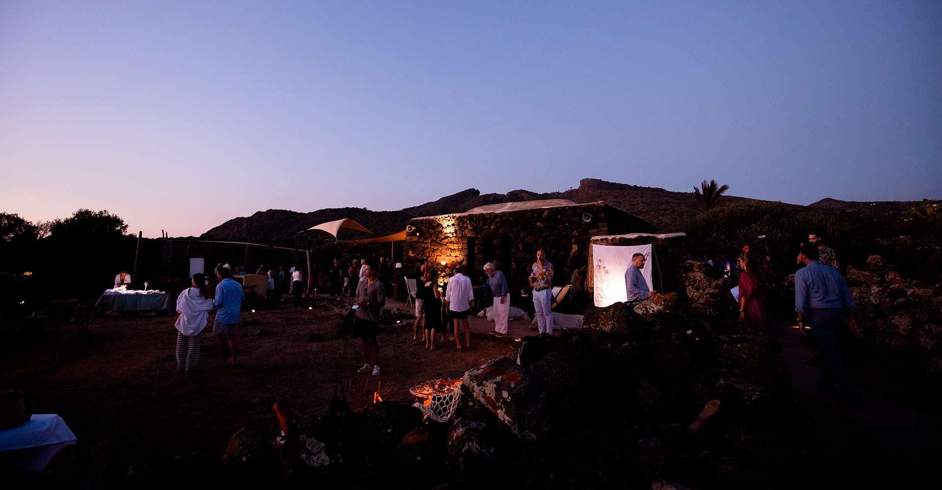 Een avondbijeenkomst buiten bij la collina di loredana, met mensen die zich vermengen bij stenen bouwwerken. Zachte verlichting creëert een warme sfeer tegen de schemerige hemel en heuvels.