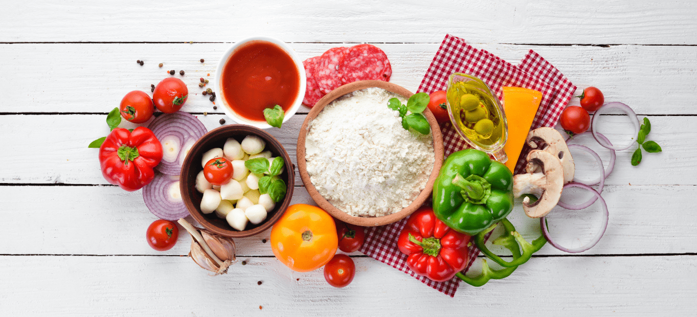 Verschillende pizza-ingrediënten op een witte houten tafel, waaronder bloem, cherrytomaten, paprika's, kaas, uien, saus en champignons, zorgen voor een fris en levendig tafereel.